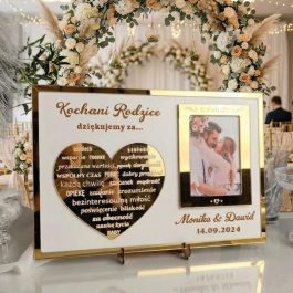 podziękowania dla rodziców w złotej ramie, ustawiona na tle eleganckiej sali weselnej z kwiatowym łukiem. Na tabliczce znajduje się zdjęcie pary młodej, złote serce z wypisanymi wartościami oraz napis „Kochani Rodzice, dziękujemy za...”.