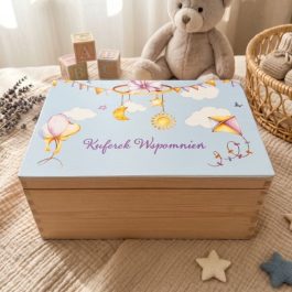Drewniany kuferek wspomnień dla dziecka z ilustracją nieba i napisem na wieku, umieszczony na niebieskim, dzierganym kocyku w przytulnym pokoju dziecięcym obok misia i książek.