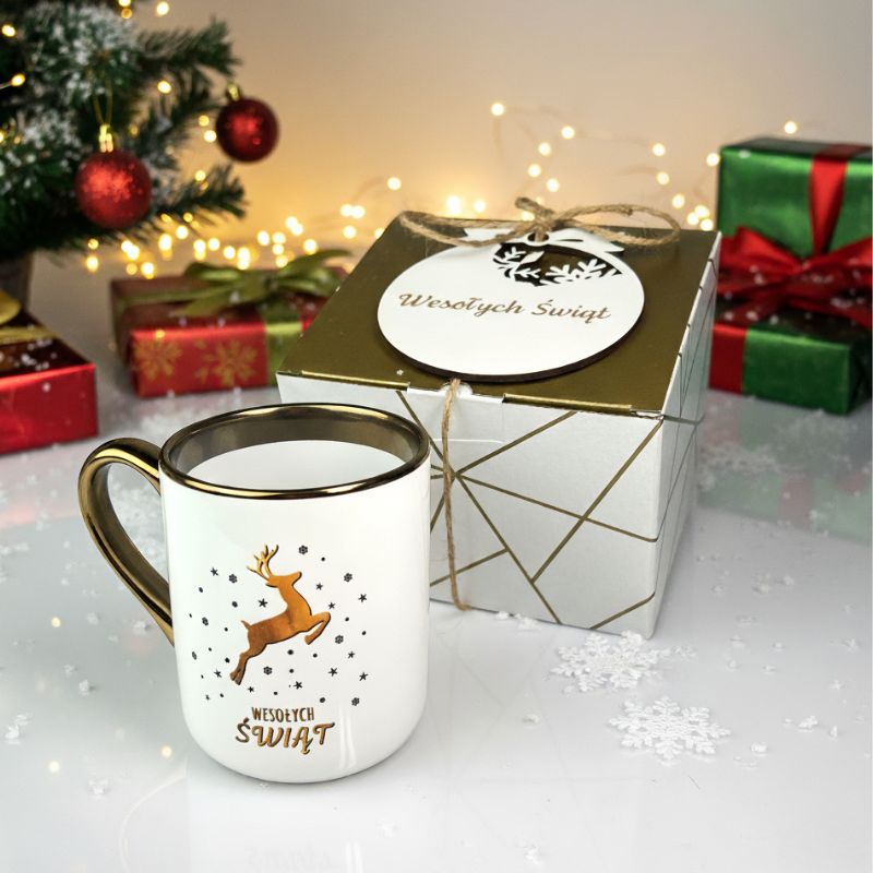 Prezent Świąteczny dla Pracowników Zestaw Kubek Świąteczny i bombka MD1650. Zestaw świąteczny z kubkiem i bombką z grawetem wesołych świąt. Kubek biały ze złotym paskiem i uchem z nadrukiem reniferka złotego.