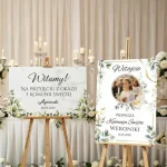 Dwie tablice komunijne. Biała tablica powitalna na Komunię Świętą ze złotym napisem na drewnianej sztaludze, dekorowana kwiatami. Druga tablica na komunię ze zdjęciem dziecka. Ustawione na sztaludze – inspiracje My-Deco