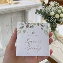 eleganckiego pudełka na pamiątkę Pierwszej Komunii Świętej o wymiarach 9x9 cm. Widoczny subtelny motyw zielonych gałązek eukaliptusa, srebrny grawer IHS oraz ozdobna kaligrafia. Pudełko prezentuje się jako luksusowe etui na różaniec lub medalik.