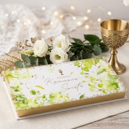 Prostokątne, eleganckie pudełko na pieniądze prezentowe na pieniądze na Pierwszą Komunię Świętą. Białe tło z delikatną złotą i zieloną grafiką roślinną oraz napisem "Pamiątka I Komunii Świętej". Pudełko jest zamknięte, widoczne pod lekkim skosem na jasnym tle.