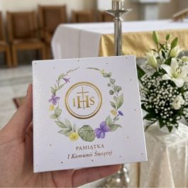 eleganckiego pudełka na pamiątkę Pierwszej Komunii Świętej o wymiarach 9x9 cm. Widoczny subtelny motyw zielonych gałązek eukaliptusa, srebrny grawer IHS oraz ozdobna kaligrafia. Pudełko prezentuje się jako luksusowe etui na różaniec lub medalik.