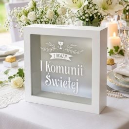 Skarbonka w formie białej ramki 15x15 cm z nadrukiem na szkle „Z okazaji I Komunii Świętej” na tle grafik zielonych liści i kielicha z hostią. Widok z perspektywy.