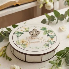 Pamiątka Pierwszej Komunii Świętej . Otwarta, okrągła drewniana szkatułka na pamiątkę Pierwszej Komunii Świętej, stojąca na białym, przecieranym stole. Wewnątrz widać metalową przywieszkę w kształcie ażurowego anioła z białym chwostem oraz błękitny różaniec z perełek. Obok leży wieczko z botanicznym wzorem wianka, złotym uchwytem i napisem "Pamiątka Pierwszej Komunii Św.", a także mała biała książeczka, koronka i płatki kwiatów.