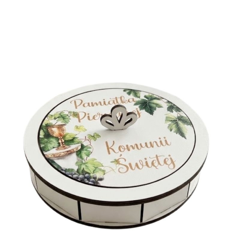 Pamiątka Pierwszej Komunii Świętej . Otwarta, okrągła drewniana szkatułka na pamiątkę Pierwszej Komunii Świętej, stojąca na białym, przecieranym stole. Wewnątrz widać metalową przywieszkę w kształcie ażurowego anioła z białym chwostem oraz błękitny różaniec z perełek. Obok leży wieczko z botanicznym wzorem wianka, złotym uchwytem i napisem "Pamiątka Pierwszej Komunii Św.", a także mała biała książeczka, koronka i płatki kwiatów.