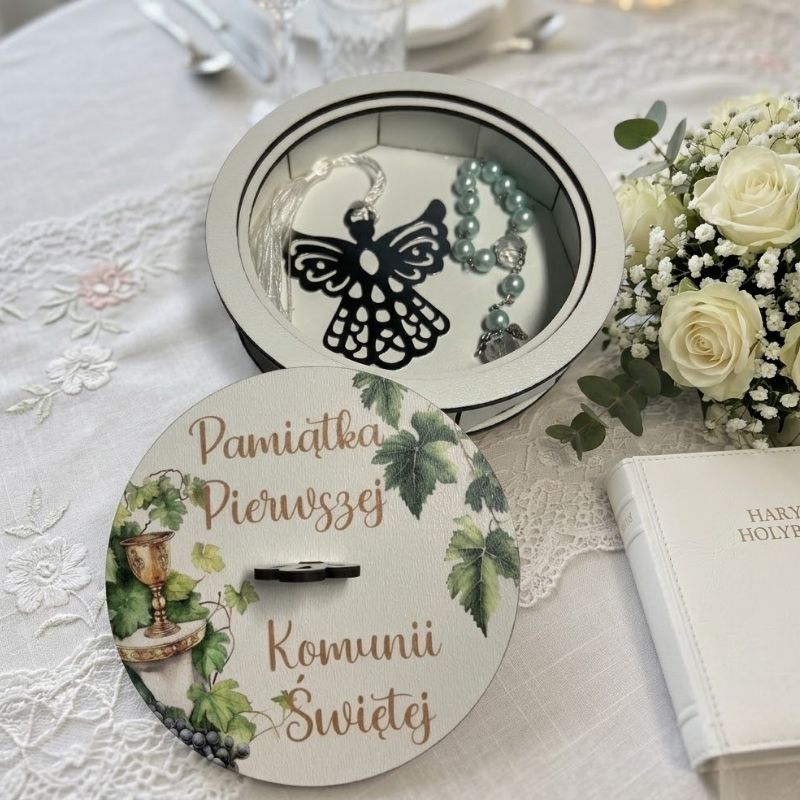 Pamiątka Pierwszej Komunii Świętej . Otwarta, okrągła drewniana szkatułka na pamiątkę Pierwszej Komunii Świętej, stojąca na białym, przecieranym stole. Wewnątrz widać metalową przywieszkę w kształcie ażurowego anioła z białym chwostem oraz błękitny różaniec z perełek. Obok leży wieczko z botanicznym wzorem wianka, złotym uchwytem i napisem "Pamiątka Pierwszej Komunii Św.", a także mała biała książeczka, koronka i płatki kwiatów.