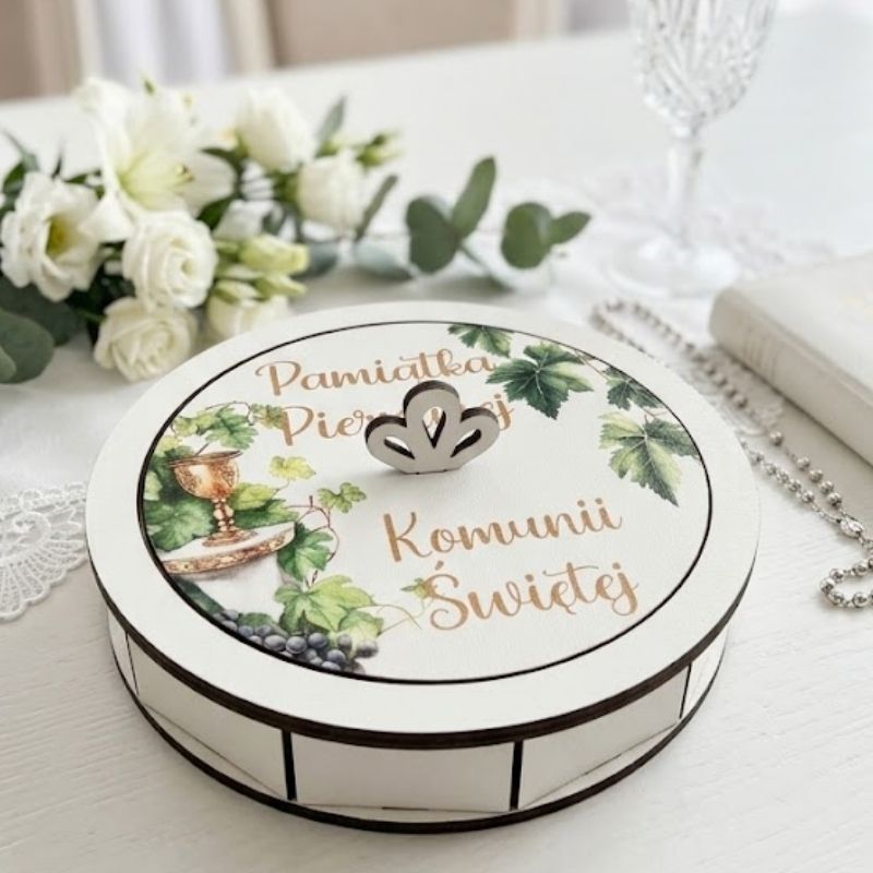 Białe pudełko, Pamiątka Pierwszej Komunii Świętej . Otwarta, okrągła drewniana szkatułka na pamiątkę Pierwszej Komunii Świętej, stojąca na białym, przecieranym stole. Wewnątrz widać metalową przywieszkę w kształcie ażurowego anioła z białym chwostem oraz błękitny różaniec z perełek. Obok leży wieczko z botanicznym wzorem wianka, złotym uchwytem i napisem "Pamiątka Pierwszej Komunii Św.", a także mała biała książeczka, koronka i płatki kwiatów.