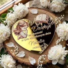 Upominek dla gości. Personalizowany magnes na podziękowanie komunijne w kształcie serca ze złotej pleksi lustrzanej, z kolorowym nadrukiem modlącej się dziewczynki w wianku, otwartą Biblią, ozdobnym nadrukiem i Polish text "Dziękuję za obecność w tym wyjątkowym dniu Natalia 17.05.2027", położony na plaster drewna i otoczony białymi peoniami i gipsówką, w jasnym świetle dziennym.